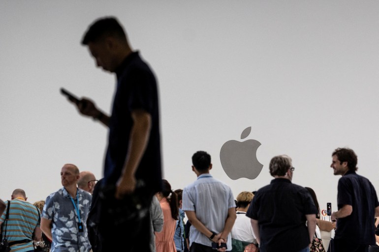 A man in the foreground stares into a phone, while others gather near an Apple logo sign